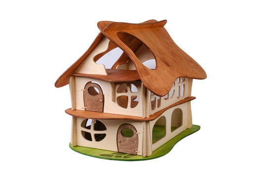 A two-story wooden dollhouse with a brown slotted roof, beige walls, and green accents representing grass at the base. Designed for children, the dollhouse features multiple windows and doors.