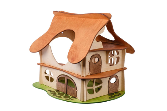 A two-story wooden dollhouse with a brown slotted roof, beige walls, and green accents representing grass at the base. Designed for children, the dollhouse features multiple windows and doors.