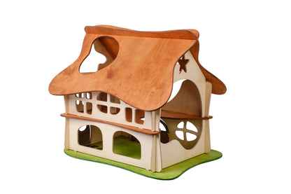 A two-story wooden dollhouse with a brown slotted roof, beige walls, and green accents representing grass at the base. Designed for children, the dollhouse features multiple windows and doors.