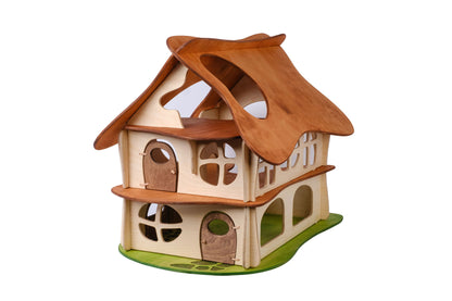 A two-story wooden dollhouse with a brown slotted roof, beige walls, and green accents representing grass at the base. Designed for children, the dollhouse features multiple windows and doors.